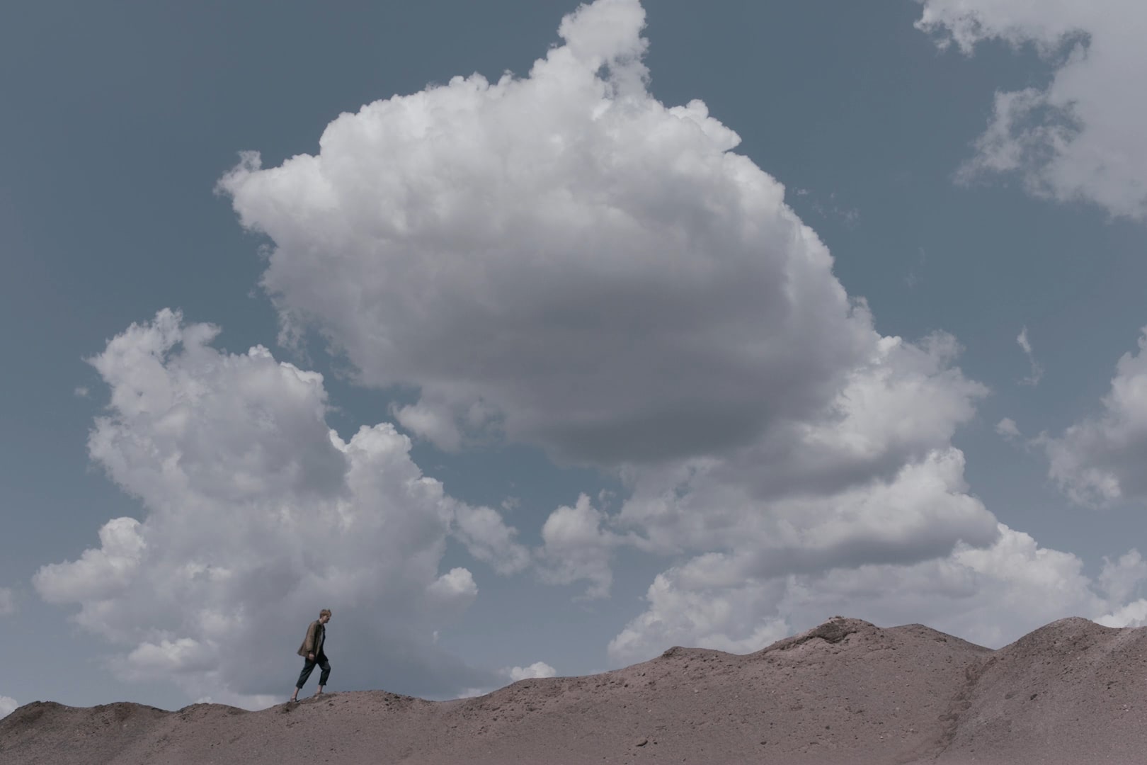 Man walking on a sand dune against the horizon beneath giant white clouds.