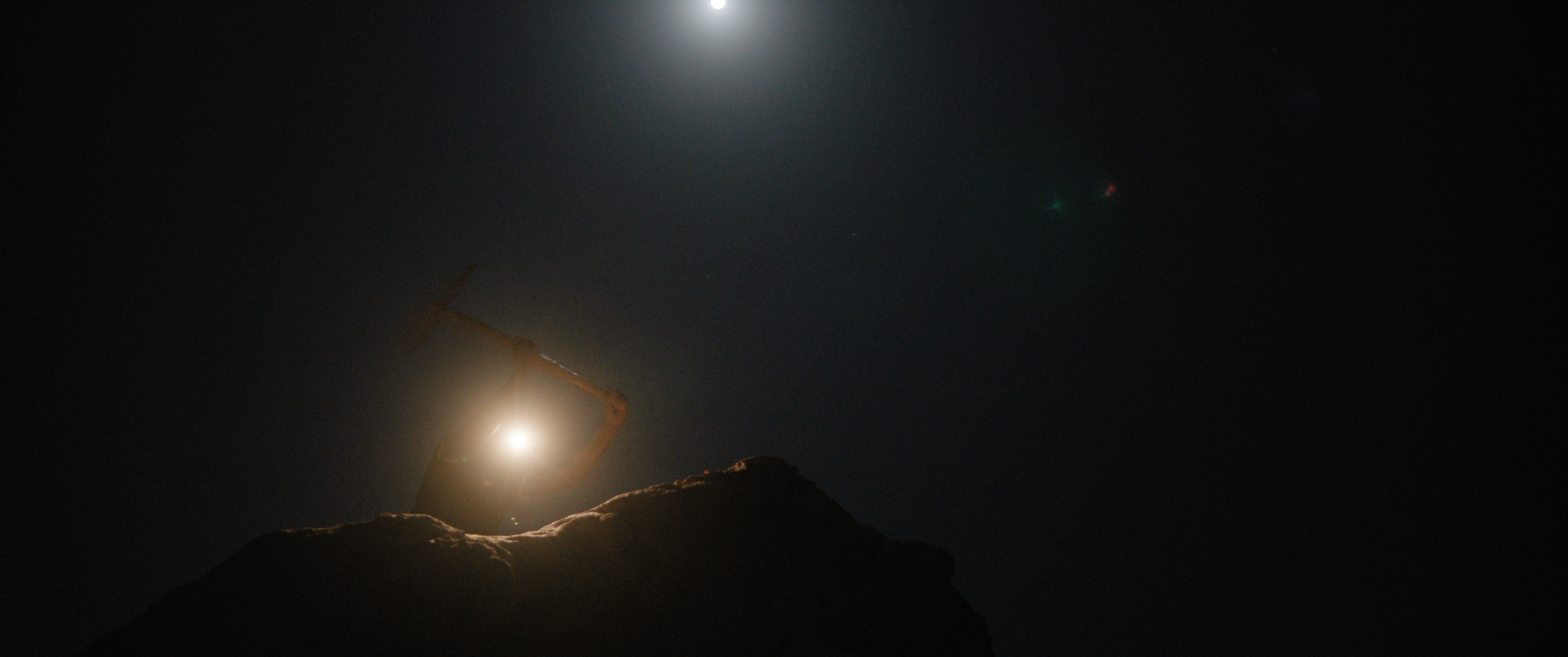 A woman at night digging with a pick axe and a headlamp, with the moon behind her.