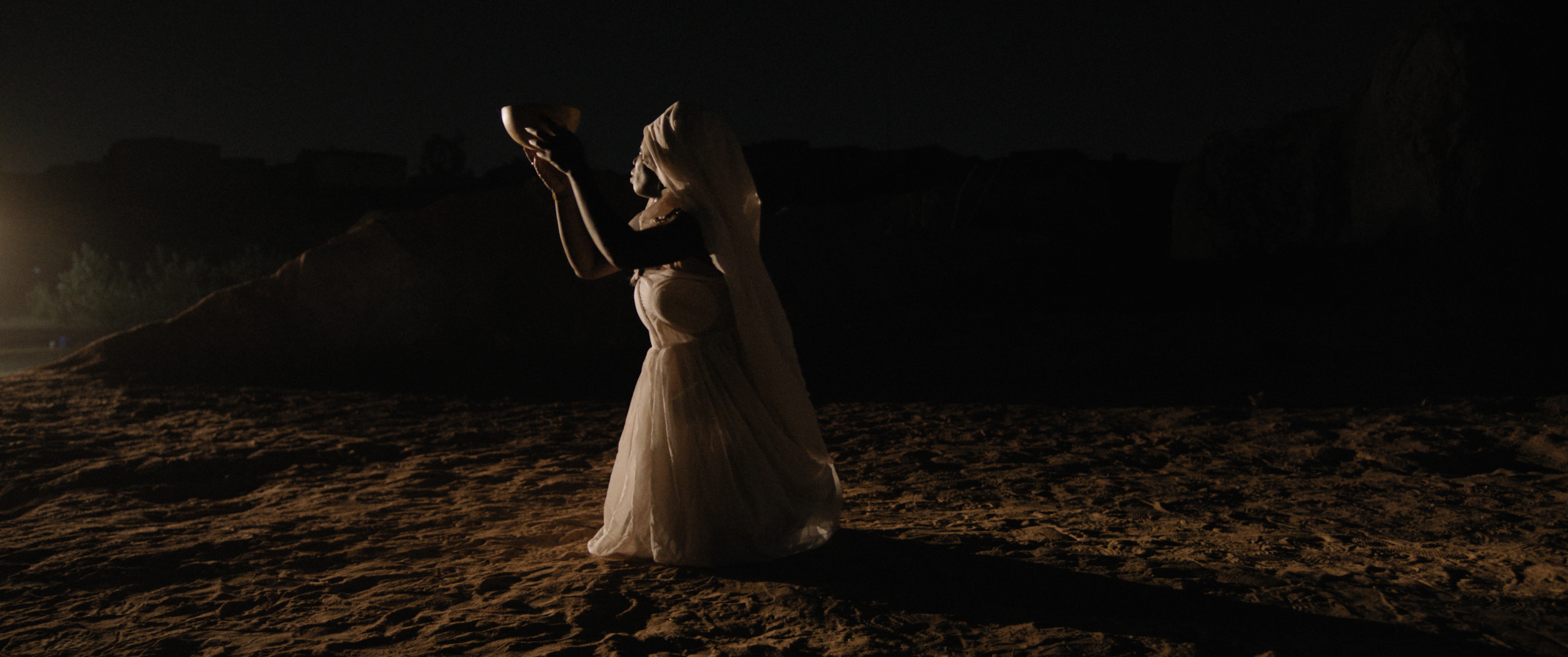 A woman in a white dress at night, kneeling in the sand and holding a bowl