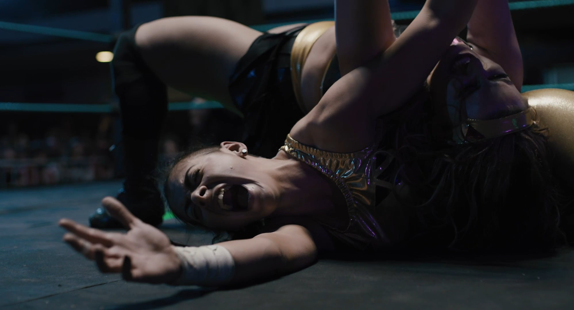 A young woman lying on the floor of the ring, screaming in pain, while another woman is on top of her.