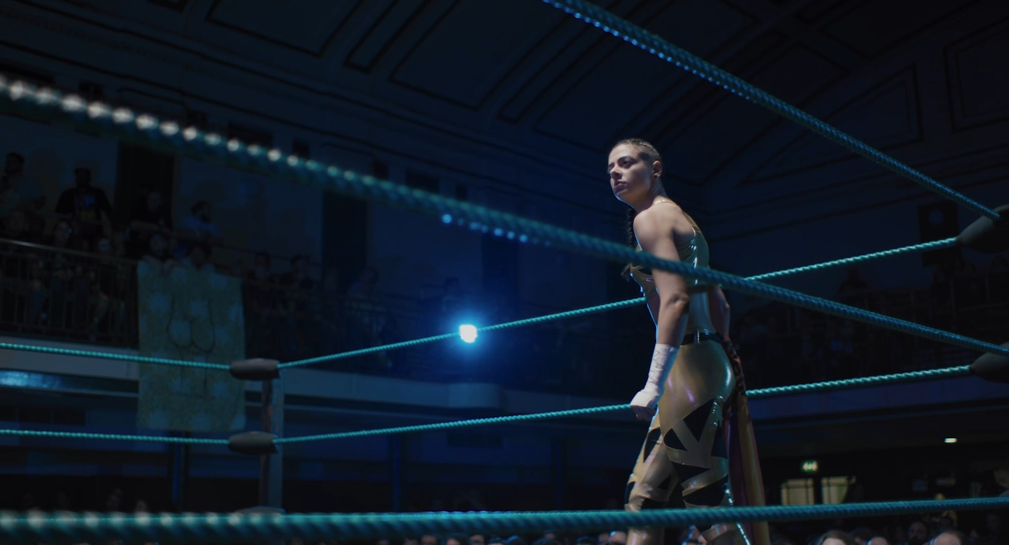 A young woman in a gold costume standing inside a wrestling ring with a large crowd watching.