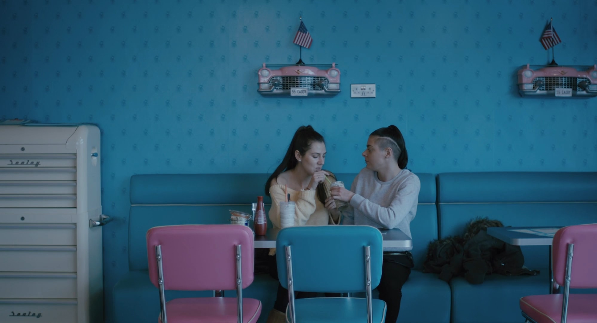 Two young women in an American diner drinking a milk shake and looking at each other.