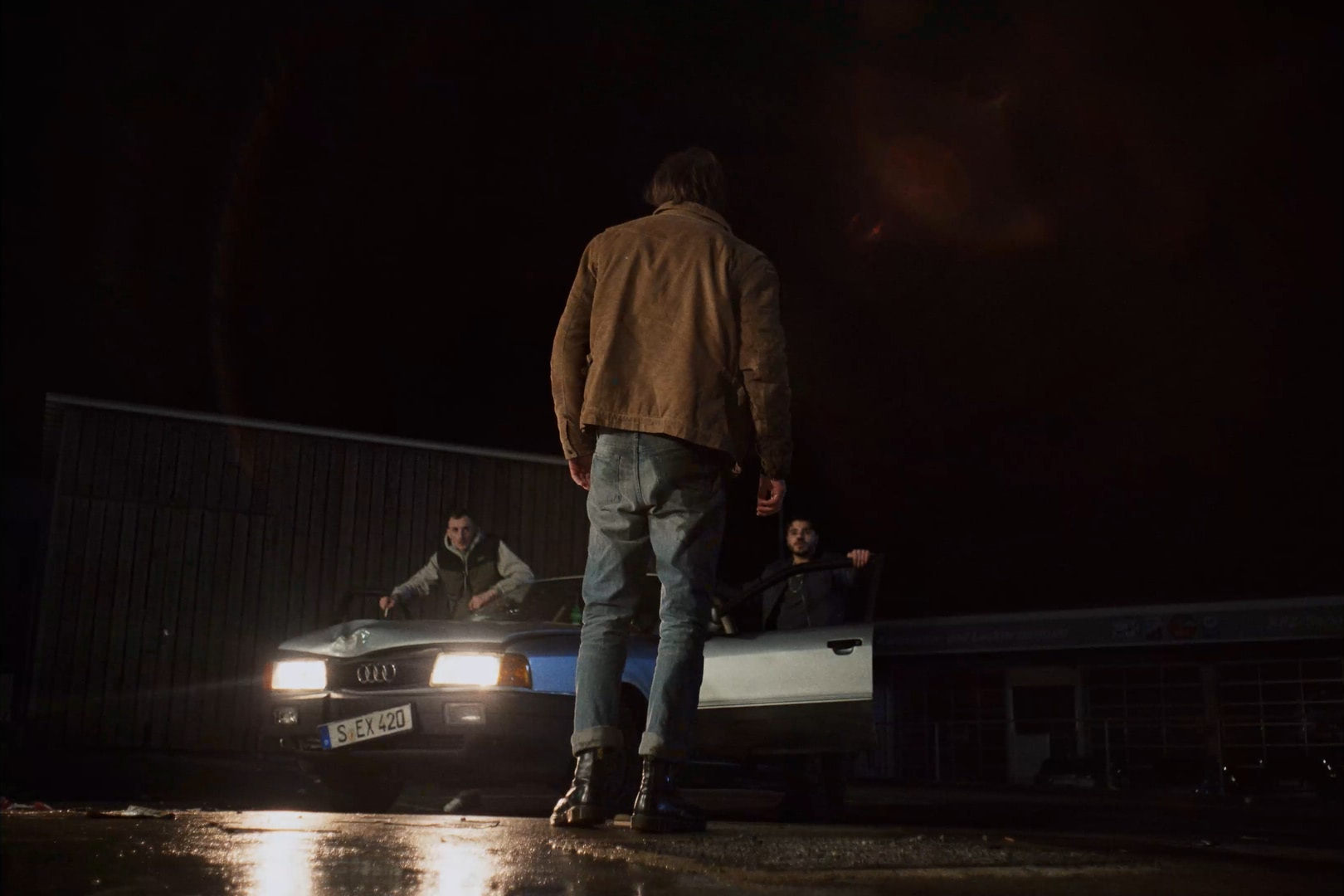 A man from behind facing the lights of a car, two other men are facing him.