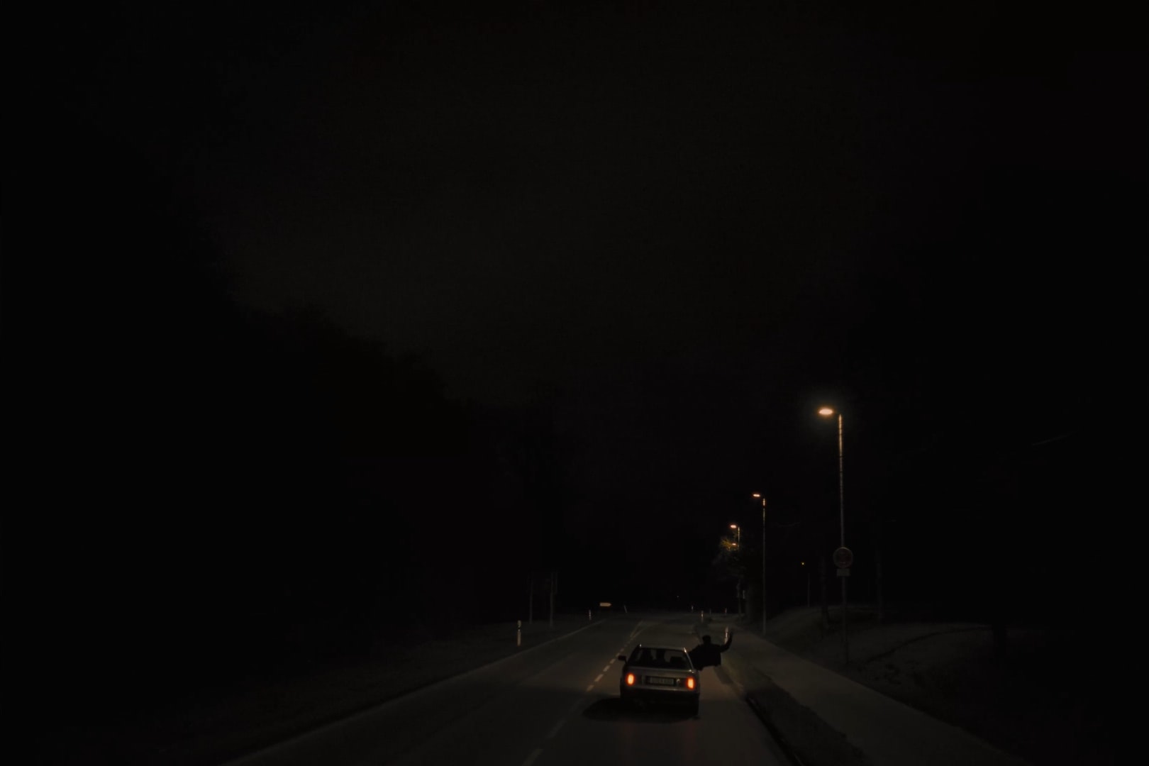 Wide shot of a car driving a small road at night and a figure leaning out the window.