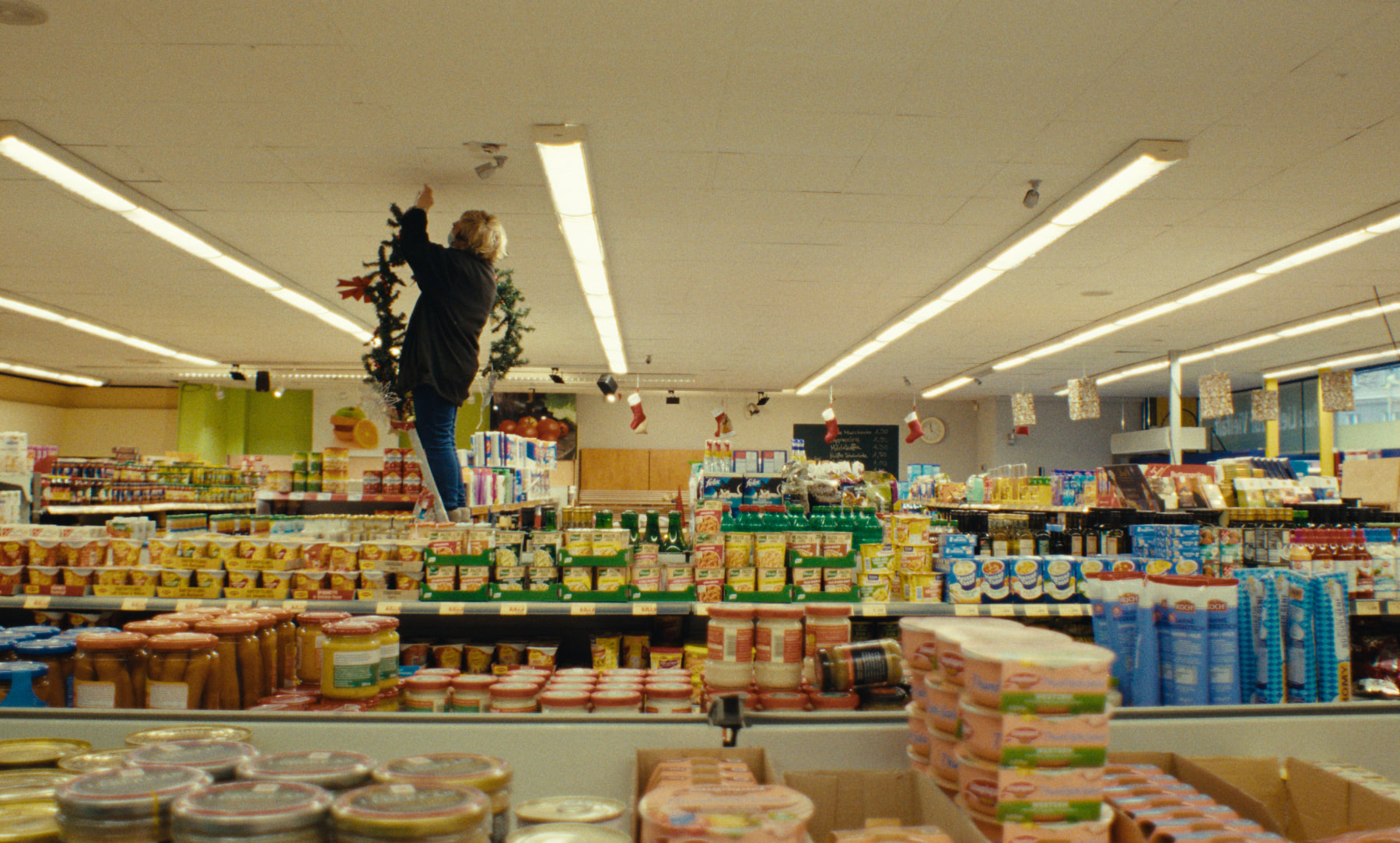 A woman standing on a ladder between supermarket aisles, attaching holiday decoration to the ceiling. 