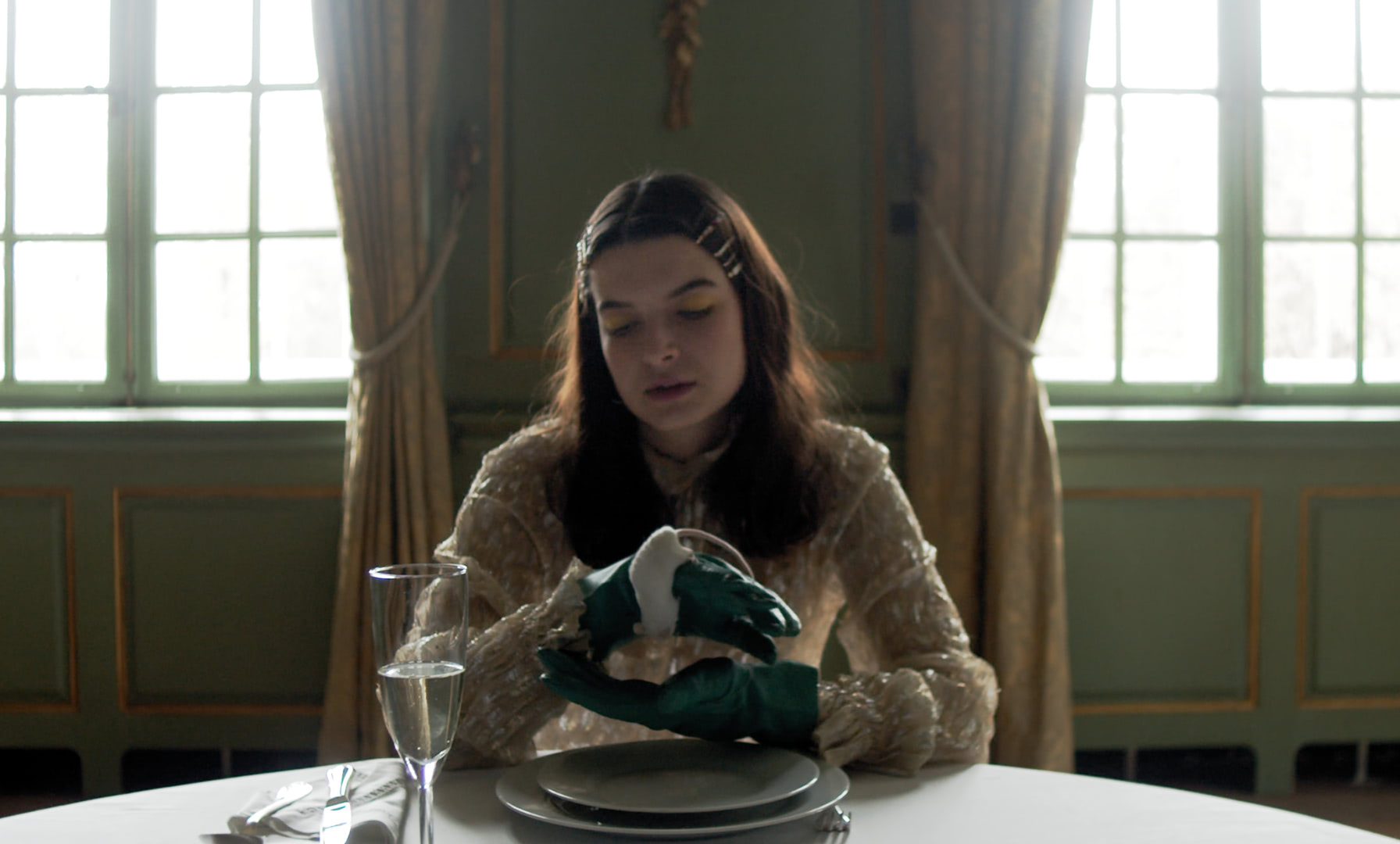 A young woman sitting at the table of an opulent green dining room, holding a white mouse in her hand.