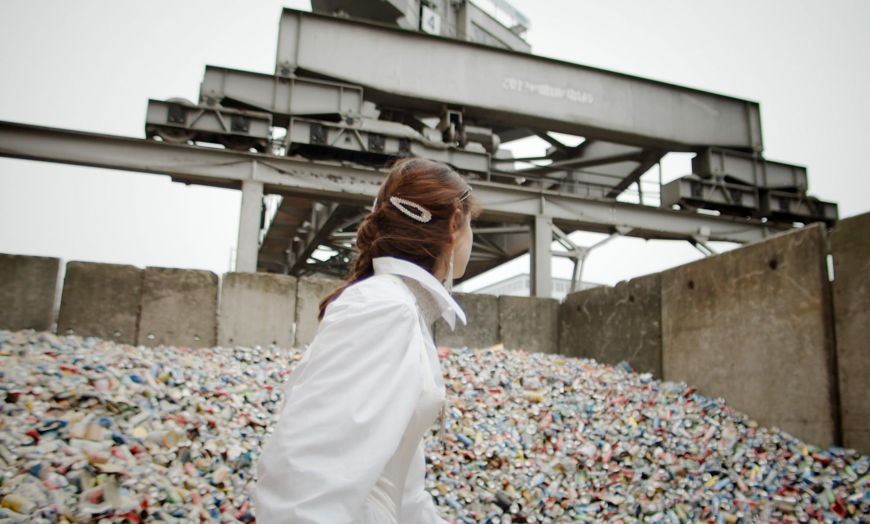 A young woman is standing in a recycling yard in front of a mountain of old discarded beer cans.