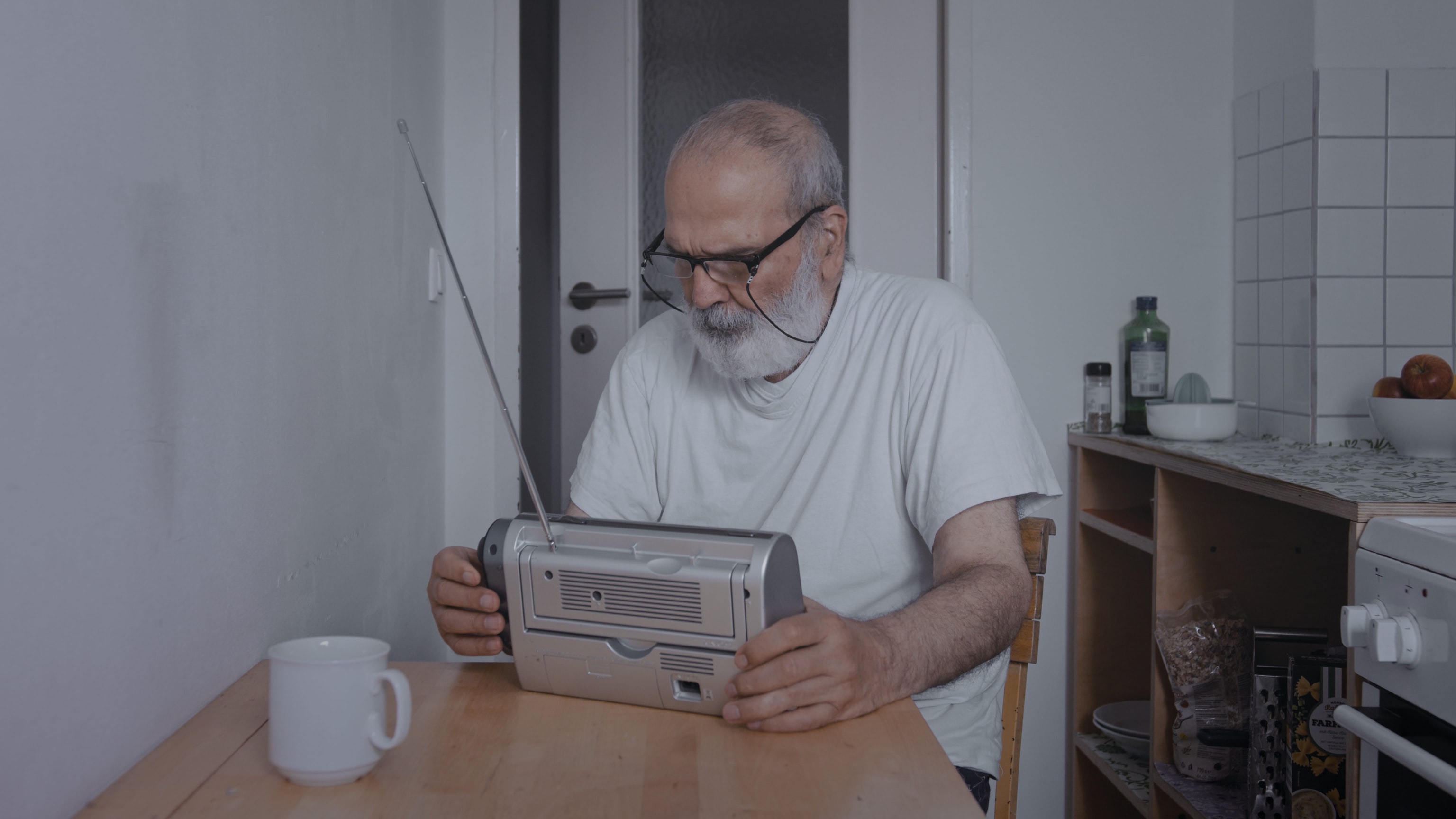An older middle eastern sitting in the kitchen, tuning a small transistor radio,