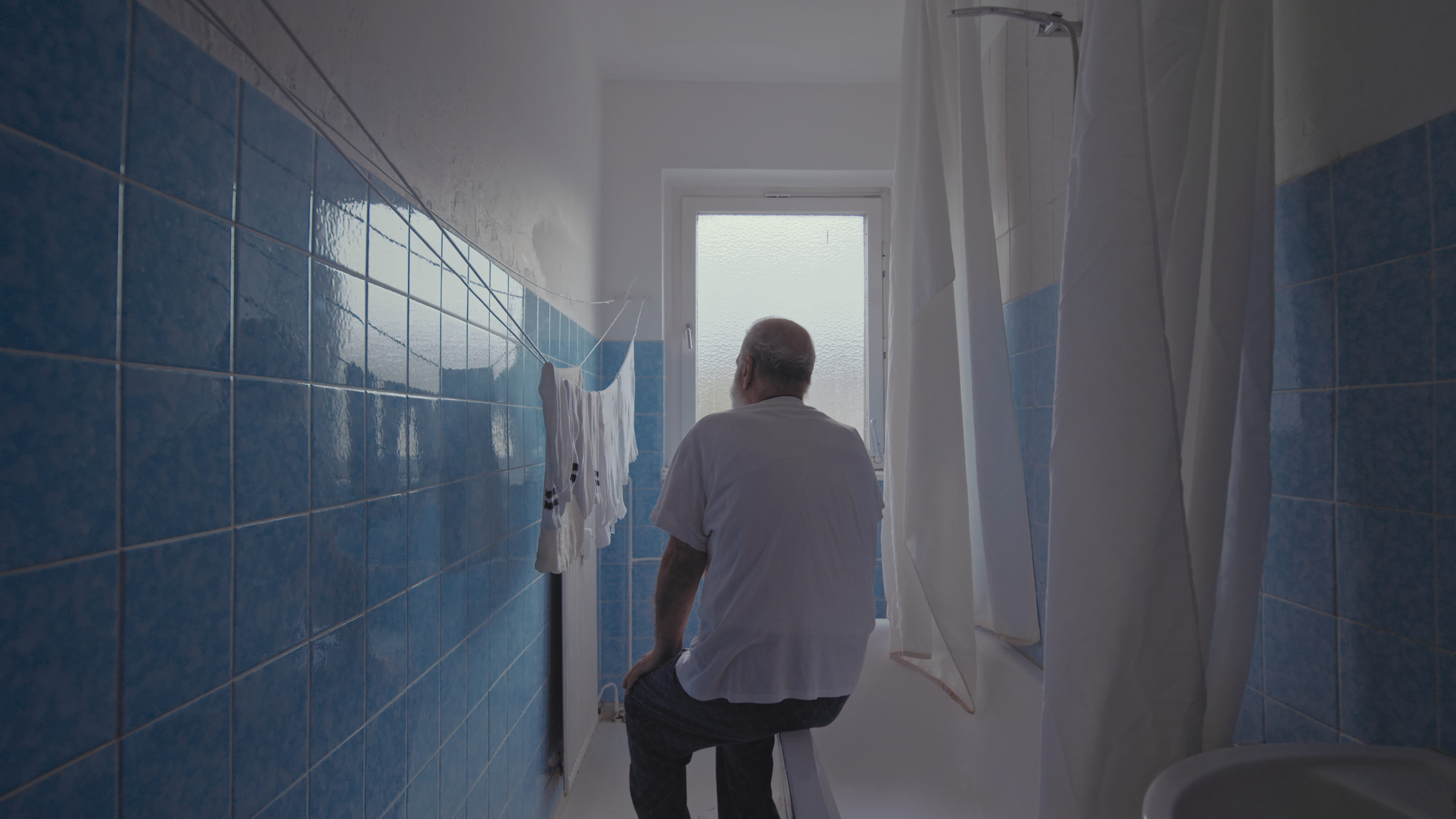 An older middle eastern man sitting on the edge of the bath tub in a bathroom with blue tiles.
