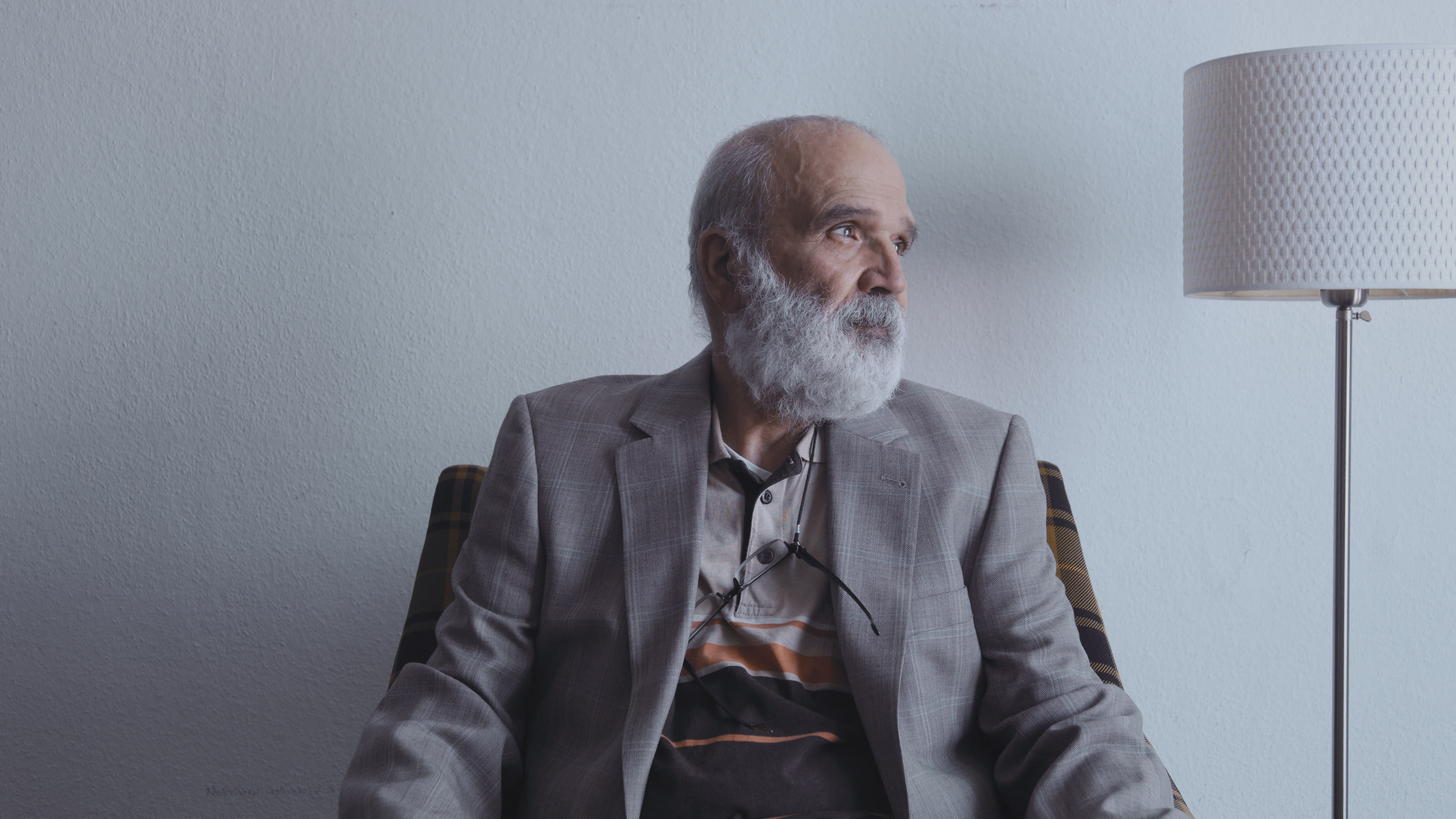 An older Middle Eastern man sitting on an arm chair in front of a white wall. 