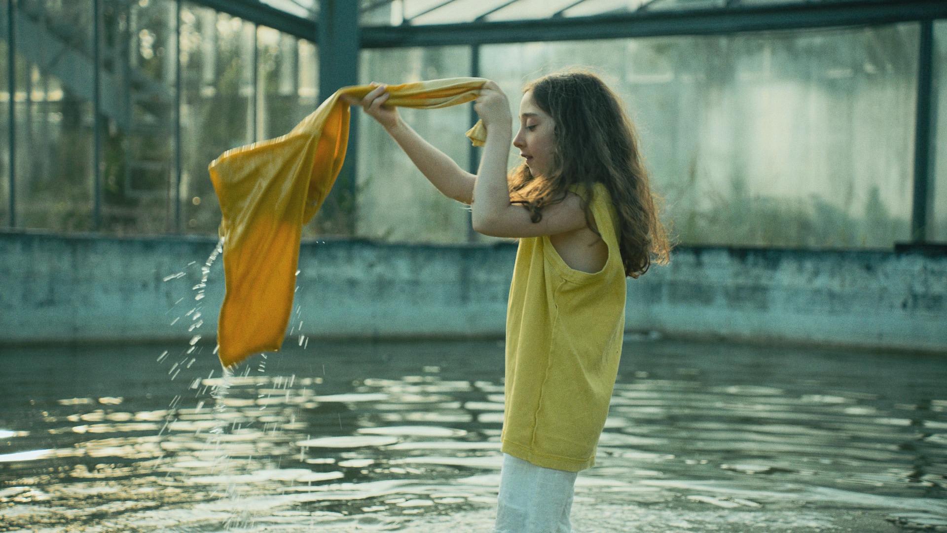 A girl standing in a pool wrings the water out of a yellow piece of wet clothing.